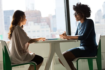 Two women sitting and talking at a table with a city view from the window. HD wallpaper