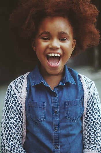 A cheerful young girl with afro hair wearing a denim shirt smiling outdoors. HD phone wallpaper