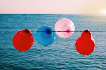 Vibrant red, blue, and pink balloons hanging on a seaside line during sunset. HD wallpaper