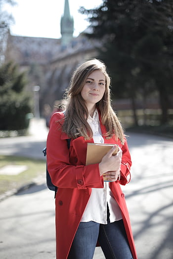 Young woman in a red coat smiling while holding a book outside on a sunny day. HD phone wallpaper