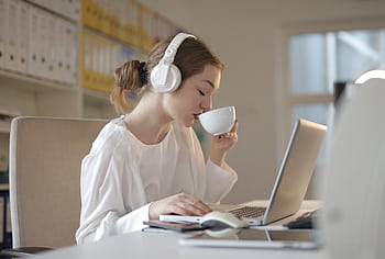 Woman wearing heads and sipping coffee while working on a laptop at a desk. HD wallpaper