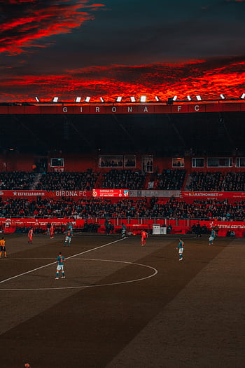 Dramatic evening soccer game at Girona FC stadium with vibrant red sky and intense play. HD phone wallpaper
