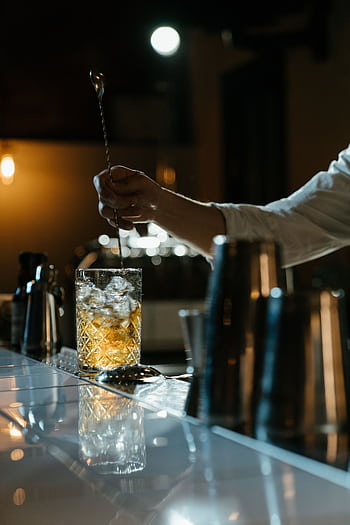 A bartender stirring a whiskey cocktail with a spoon in a dimly lit bar setting. HD phone wallpaper