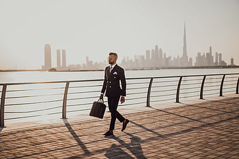 Professional man in formal suit walking by Dubai waterfront with city skyline and Burj Khalifa in view. HD wallpaper