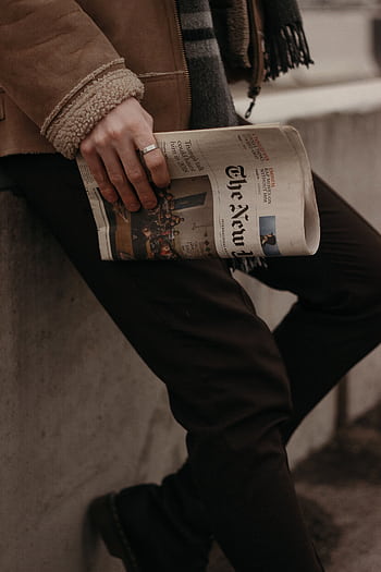 A fashionable man holds a newspaper while leaning against a city wall, showcasing modern urban style. HD phone wallpaper