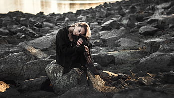 A woman in a black outfit sitting on rocks by the water, reflective mood. HD wallpaper