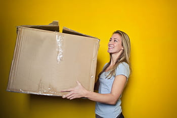 A woman carrying a large cardboard box against a vibrant yellow background, smiling. HD wallpaper