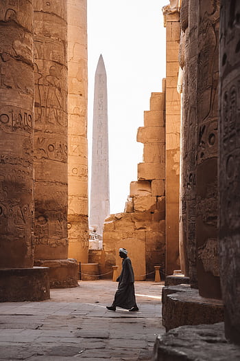 A serene view of the Karnak Temple's ancient columns and obelisk with a person walking through. HD phone wallpaper