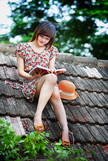 Cheerful young woman in a floral dress reading a book on a rooftop, enjoying a sunny day. HD phone wallpaper
