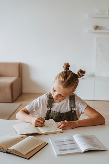 A young girl sits at a table indoors, engaged in writing in a notebook with open books around her, embodying a focused study environment. HD phone wallpaper