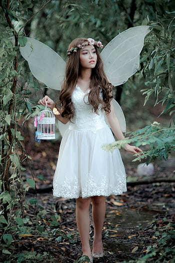Fairy in mystical forest wearing a white dress and wings, holding a decorative birdcage. HD phone wallpaper