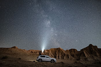 A person stands on an SUV under a vast starry night sky in the desert, amid rock formations. HD wallpaper