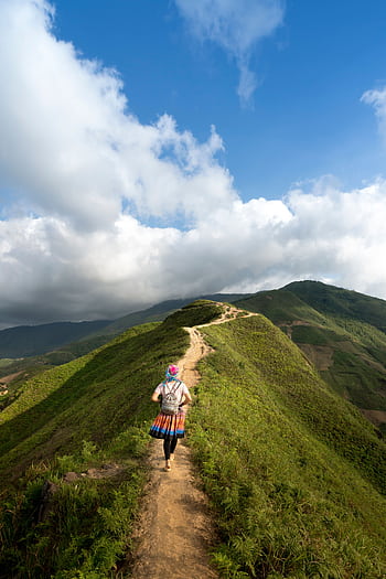 A lone hiker walking on a scenic mountain trail surrounded by lush greenery and clear blue skies. HD phone wallpaper