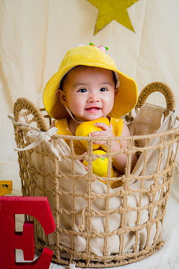 Cute baby in yellow hat smiling joyfully inside a woven basket with toys. HD phone wallpaper