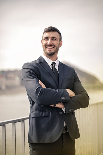 Smiling businessman in a black suit standing outdoors by a railing, showcasing a confident demeanor. HD phone wallpaper