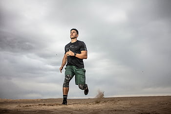 A muscular man running on sandy terrain under cloudy skies, promoting fitness and adventure. HD wallpaper