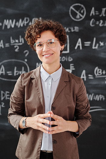 Teacher standing by a blackboard with algebra equations, smiling confidently. HD phone wallpaper