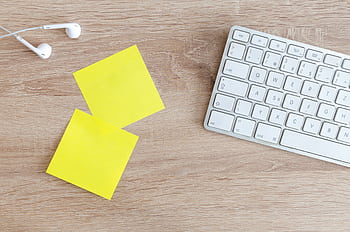 Top view of a workspace with a keyboard, ears, and yellow sticky notes on a wooden desk. HD wallpaper