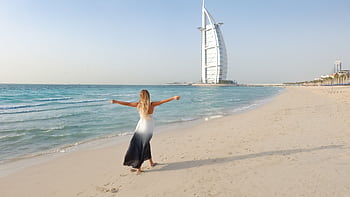 A care woman in a flowing dress strolls along the Dubai beach with Burj Al Arab in view. HD wallpaper