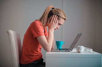 A woman sits indoors at a table, looking concerned while using a laptop with heads and a coffee mug nearby. HD wallpaper