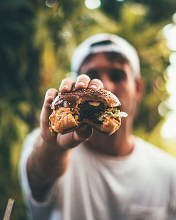 A man holds a juicy cheeseburger outdoors in daylight, highlighting its deliciousness and freshness. HD phone wallpaper