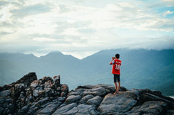 A person enjoying a scenic view on a rocky mountain peak, surrounded by lush landscapes and wide skies. HD wallpaper