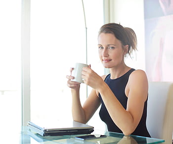 Young woman sitting at her desk, holding a coffee cup and smiling. Ideal for workplace and lifestyle themes. HD wallpaper
