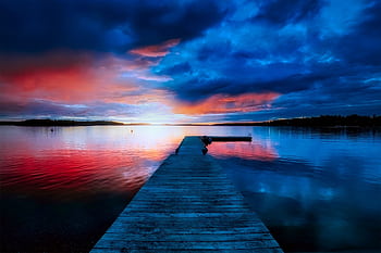 A tranquil sunset view with vibrant hues reflecting over a calm lake and a wooden dock extending into the water. HD wallpaper