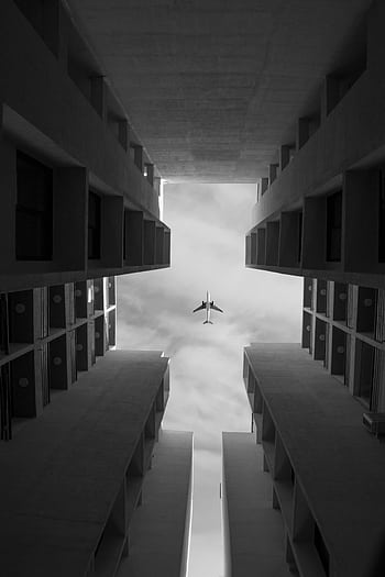 Low angle of symmetrical buildings against sky with airplane. Monochrome. HD phone wallpaper