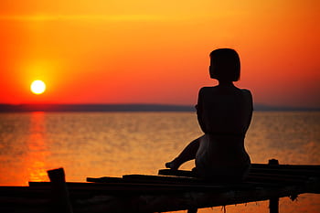 Serene silhouette of a woman sitting on a pier, watching the ocean at sunset. HD wallpaper