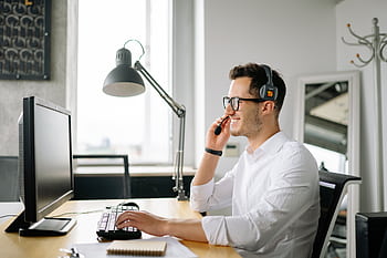 Smiling man wearing headset working at computer in bright office. HD wallpaper