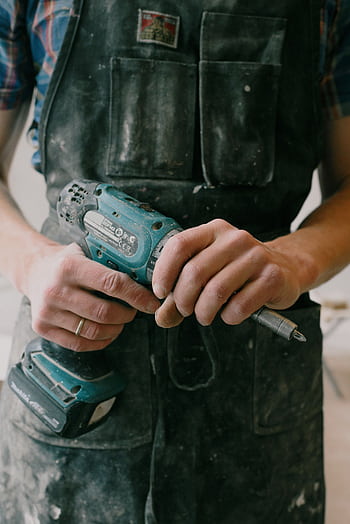 Hands of a man in workwear holding a wireless power drill indoors, capturing a carpentry moment. HD phone wallpaper
