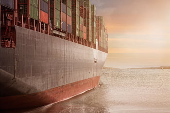 A colossal cargo ship loaded with containers navigates through calm waters against a vibrant sunset sky. HD wallpaper