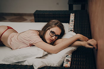 Young woman relaxing on a bed indoors, wearing glasses and casual attire. HD wallpaper