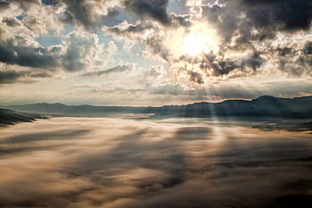 Captivating view of the sun breaking through clouds over a mist-covered mountain range. HD wallpaper