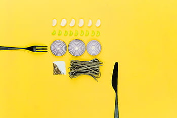 A colorful flatlay of pasta, onion, garlic, herbs, and cutlery on a yellow background. HD wallpaper