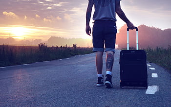 Man with tattoo walks along a rural road at sunrise with a suitcase, showcasing travel and adventure. HD wallpaper