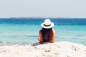 Back view of a woman in a hat relaxing on a rocky beach, facing the clear blue sea. HD wallpaper