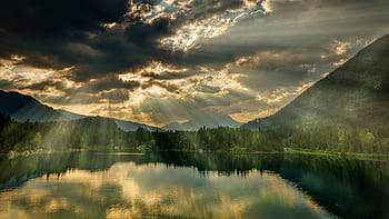 A stunning view of a tranquil lake surrounded by mountains with sunrays piercing through clouds. HD wallpaper
