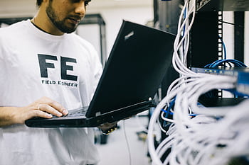 A field engineer concentrates on laptop while handling network cables in a server room. HD wallpaper