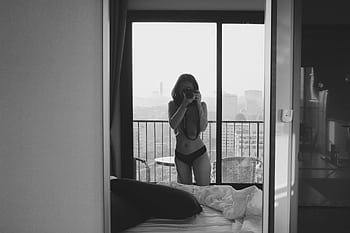 A black and white depicting a woman taking a selfie in a mirror inside a room with a city view. HD wallpaper