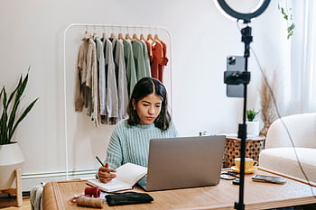Focused young woman working on laptop in home office, surrounded by clothing rack and camera setup. HD wallpaper