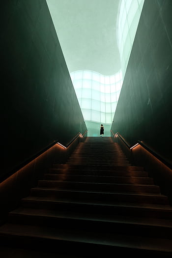A person stands at the top of a dramatic, well-lit staircase in a modern architectural interior. HD phone wallpaper