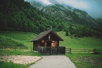 A charming wooden cabin set in a lush green valley beneath misty mountains, showcasing rural serenity. HD wallpaper