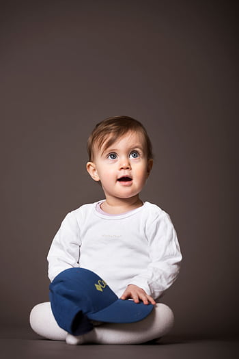 Cute baby sitting indoors, wearing white with a cap, joyful expression. HD phone wallpaper