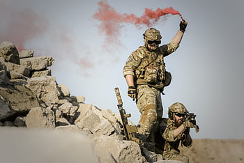 Two soldiers in camouflage, one signaling with red smoke in a desert setting. HD wallpaper
