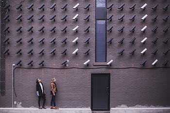 Women observing a wall covered with numerous security cameras in an urban setting, showcasing modern surveillance. HD wallpaper