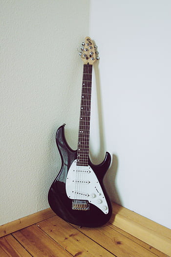 Black electric guitar with white pickguard resting against a corner wall on wooden floor. HD phone wallpaper