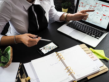A business professional working on real estate project plans using multiple devices in an office setting. HD wallpaper