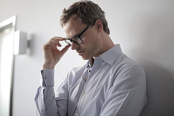 An adult man in a shirt and glasses appears stressed while leaning against a wall indoors. HD wallpaper